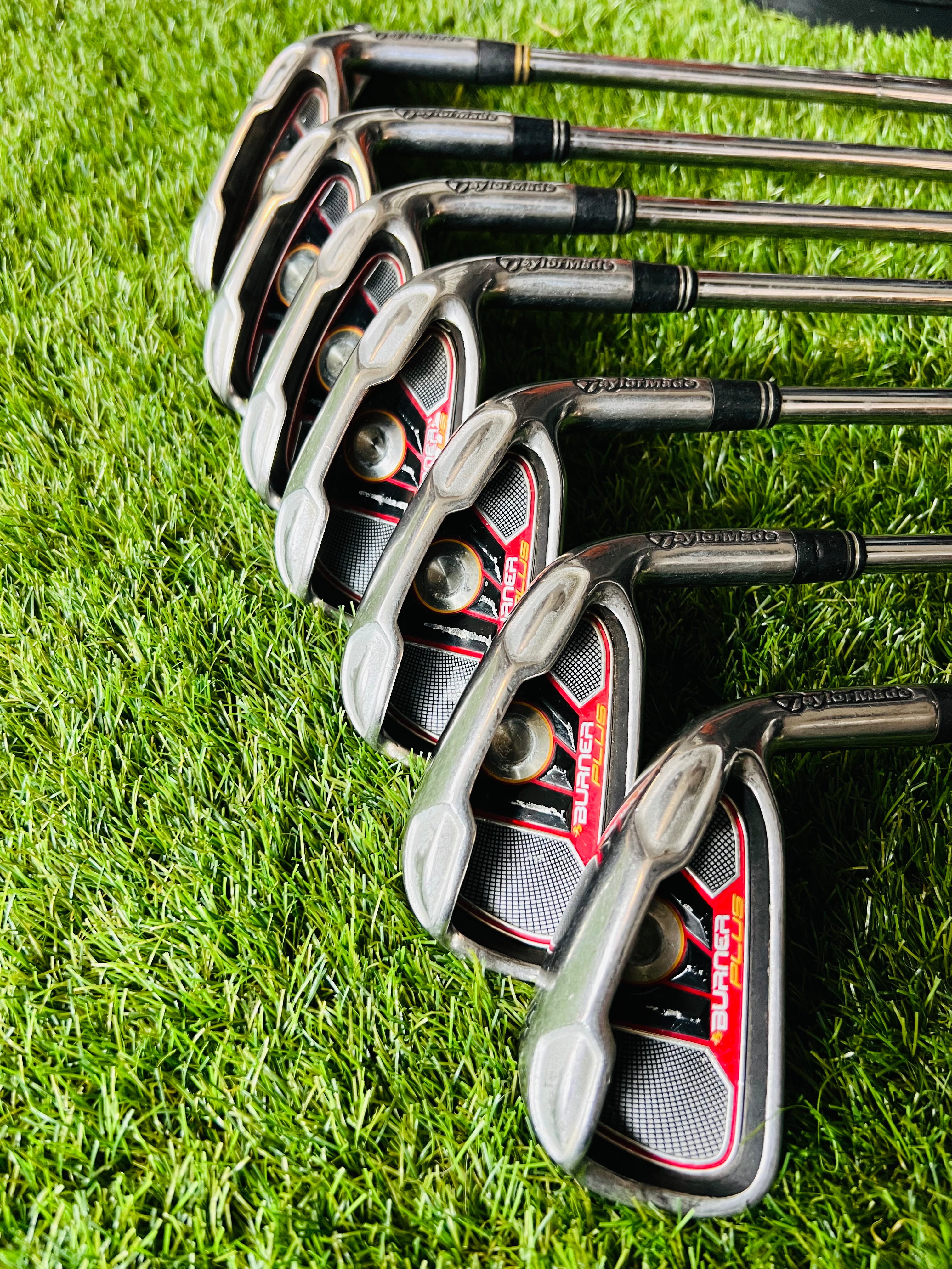 Rechtshandige TaylorMade Burner Plus ijzers 4 tot PW met +0,5 grafiet shafts, golfclubs liggen op een grasveld.