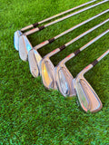 Rechtshandige Mizuno MP-20 Combo IJzers 5 tot PW liggen op een golfbaan, met houten golfclubs en golfballen op de achtergrond.